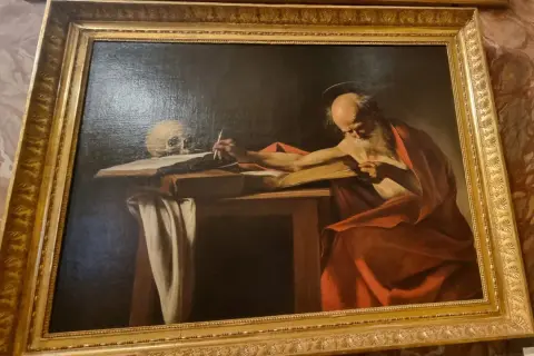 Starzec w czerwonej todze, przypominający Hieronima ze Strydonu, pochyla się nad księgą, pisząc piórem. Obok czaszka.