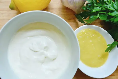 Zutaten für ein cremiges Aioli-Rezept: Knoblauch, Zitrone, Petersilie und zwei Schalen mit weißer und gelber Sauce.