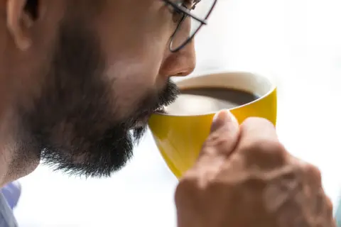 Man med glasögon och skägg dricker ur en gul mugg. Kanske känner han yrsel av kaffe.