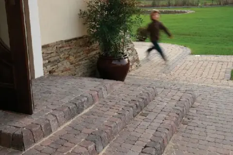 Schody z polbruku prowadzą do drzwi. W tle dziecko biegnie po trawniku.