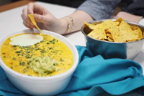 Salsa de queso cremosa con guacamole y nachos, una persona moja un totopo.