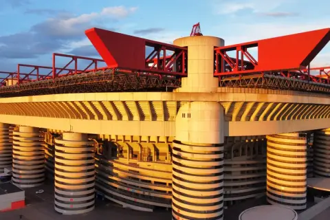 Stadion AC Milan w złotym świetle zachodzącego słońca, z charakterystycznymi czerwonymi elementami konstrukcji i betonowymi filarami.