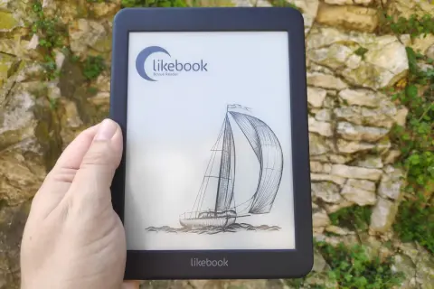 Une main tient un e-reader Likebook affichant un voilier. L'appareil est devant un mur de pierres couvert de végétation.