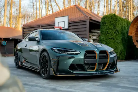 Matowy, zielony BMW M4 G82 z pomarańczowymi akcentami, zaparkowany przed drewnianym domkiem z boiskiem do koszykówki.