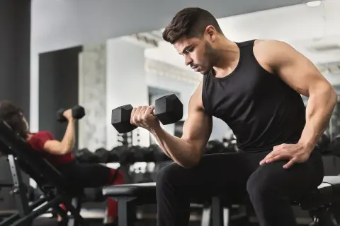 Uomo concentrato esegue esercizi con manubri in palestra. A sinistra, una donna fa lo stesso su una panca inclinata.