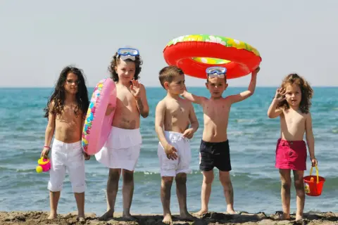 Piątka dzieci cieszy się wakacjami, gotowa na zabawy na plaży. Wśród nich są koła ratunkowe i wiaderko.