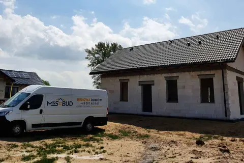 Biały van budowlany M&S BUD przed niedokończonym domem z szarej cegły pod czarnym dachem.
