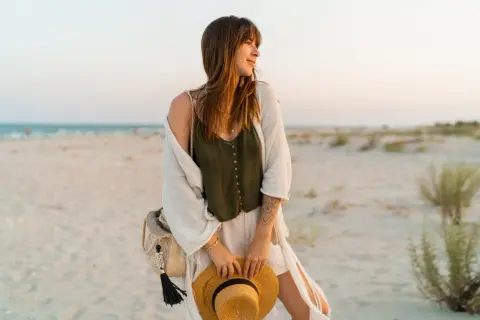 Kobieta w letnich stylizacjach boho jesień na plaży, z kapeluszem w dłoni i plecioną torbą.