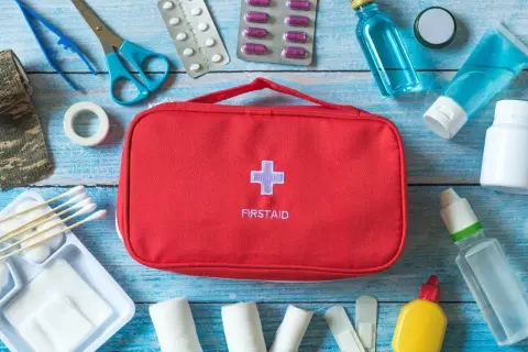 Czerwona apteczka z krzyżem i napisem FIRSTAID otoczona lekami, bandażami i akcesoriami medycznymi.