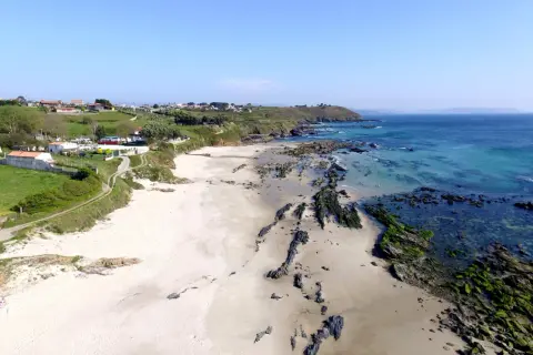 Vista aérea de la playa pragueira, con arena blanca, rocas cubiertas de algas y el mar azul turquesa. Casas y colinas verdes al fondo.
