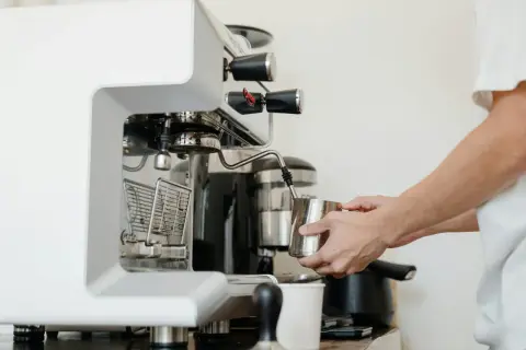 Barista spienia mleko w dzbanku, używając dyszy ekspresu. W tle widoczny blok zaparzający.