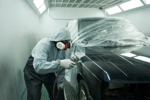 Tecnico in tuta protettiva vernicia un'auto in cabina. Il costo del ripristino trasparente dell'auto è visibile nel processo.