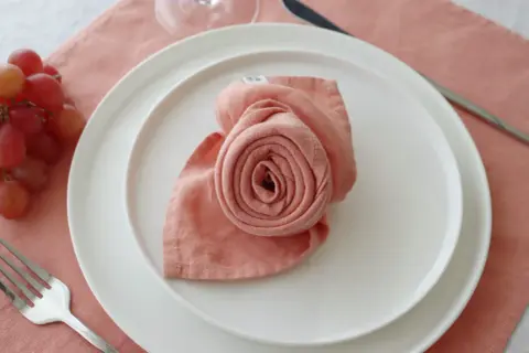 Pliage serviette rose en forme de rose sur une assiette blanche, avec une fourchette à gauche et des raisins à côté.