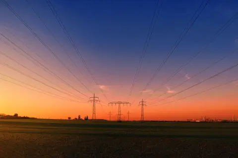 Linie energetyczne biegnące nad polem przy zachodzie słońca. W oddali widać przemysłowy krajobraz, przypominający o opłacie sieciowej stałej.