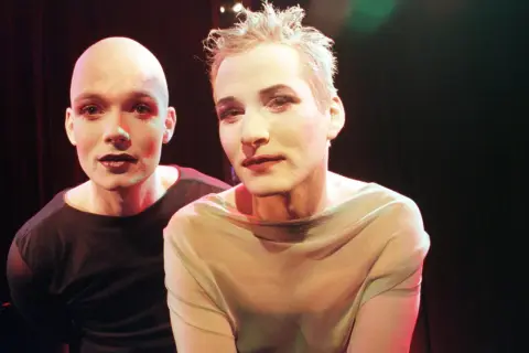 Zwei Personen mit Make-up und dunklem Lippenstift, die androgyn wirken. Eine Person ist kahlrasiert, die andere hat kurze, zerzauste Haare.