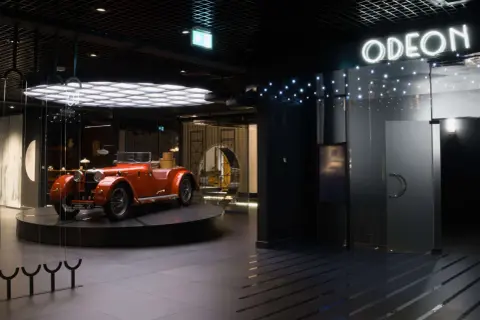 Pomarańczowy kabriolet z lat 20. XX wieku, symbol sztuki dwudziestolecia międzywojennego, prezentowany w nowoczesnym wnętrzu z neonowym napisem "ODEON".
