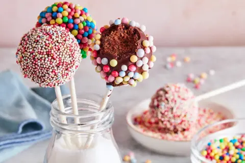 Bunte cake pops, verziert mit Zuckerstreuseln, warten darauf, vernascht zu werden. Einer ist angeknabbert und zeigt den schokoladigen Kern.
