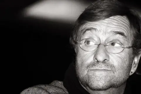 Ritratto in bianco e nero di un uomo con occhiali rotondi, barba e un'espressione pensierosa. Il suo sguardo è rivolto di lato, come se stesse ascoltando attentamente il **caruso testo**.