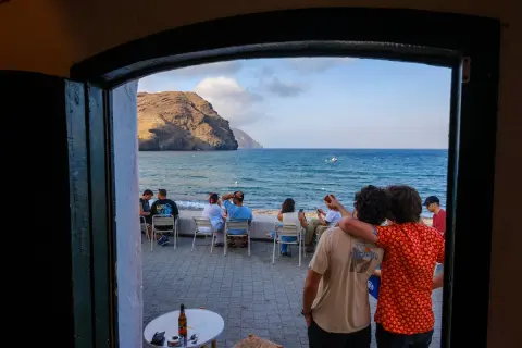 Dos amigos disfrutan de la vista al mar, con gente sentada en mesas. El manteca las negras, un día perfecto.