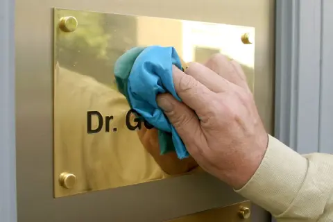 Hand reinigt Messing-Namensschild mit Tuch. Der Schriftzug "Dr. G." wird sichtbar.