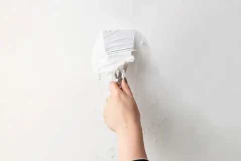 Mano con spatola che rimuove carta da parati bianca da un muro.