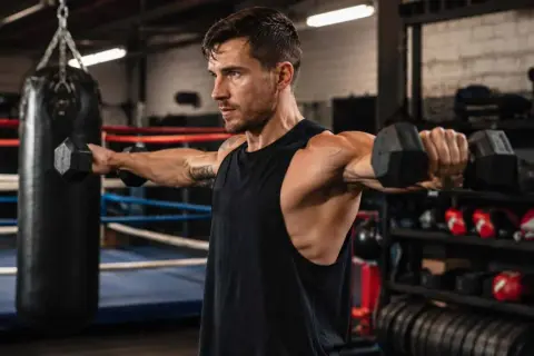 A man with impressive boxer shoulders performs dumbbell lateral raises in a boxing gym, with a punching bag in the background.