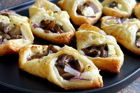 Chrupiące ciasto francuskie z pieczarkami, serem feta i czerwoną cebulą, ułożone na czarnej tacy.