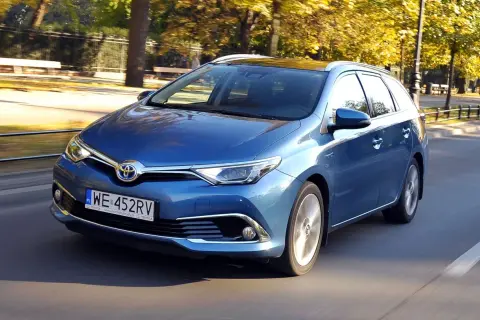 Niebieski Toyota Auris 1.8 VVT-i Hybrid w wersji kombi jedzie po drodze w otoczeniu jesiennych drzew.
