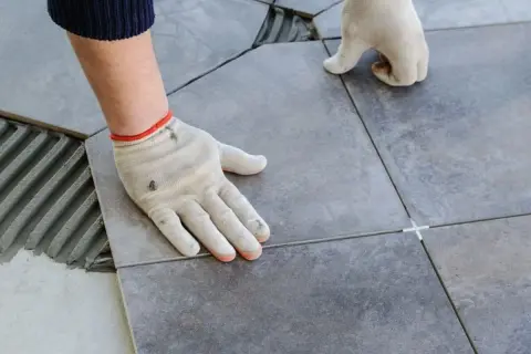 Pose de carrelage avec double encollage. Mains gantées ajustant une dalle sur un lit de mortier frais.