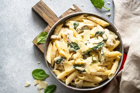 Penne arrabiata z kurczakiem i szpinakiem, posypane parmezanem, podane w patelni na drewnianej desce.