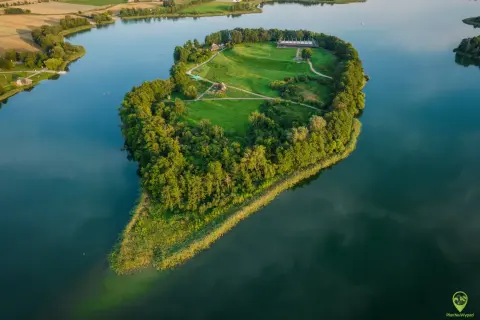 Wielkopolskie miasto koło Kalisza, Ostrowo Lednickie, z wyspą na jeziorze, porośniętą drzewami i trawą, z zabudowaniami.