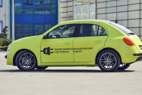 Jasnozielony FSO Syrena S106 EV, jeden z polskich samochodów elektrycznych, parkuje przed nowoczesnym budynkiem.