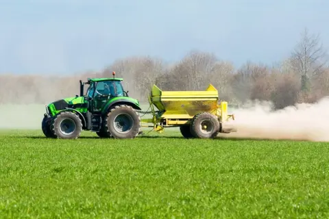 Tractor verde esparciendo cal viva en la huerta, creando una nube de polvo sobre el campo verde.