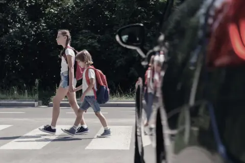 Dwoje dzieci, pieszy na pasach, z plecakami przechodzi przez ulicę.