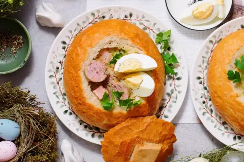 Pyszny żur w chlebie z kiełbasą i jajkiem, ozdobiony natką pietruszki. Na stole wielkanocne dekoracje: pisanki w gnieździe, zajączek.