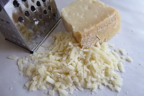 Ein Stück Käse neben einem Metallreibe mit geriebenem Käse.