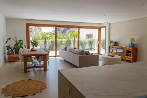 Salón moderno con comedor y sofá, destacando una pared prefabricada interior que se integra con grandes ventanales al jardín.