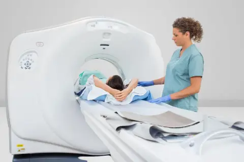 Pacjent leży na stole tomografu, pielęgniarka w rękawiczkach przygotowuje badanie PET.