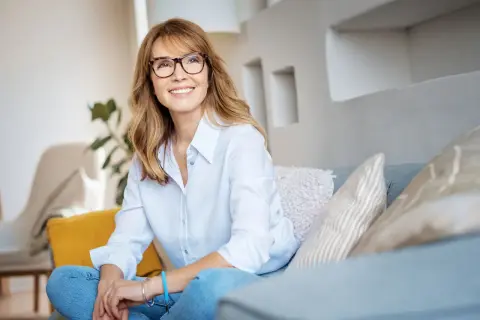 Uśmiechnięta kobieta w okularach siedzi na kanapie, zrelaksowana. Jej postawa i wyraz twarzy sugerują wysoką samoocenę, jakby wypełniała skalę samooceny Rosenberga na maksa.