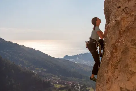 Eine Frau klettert an einer Felswand in Finale Ligure. Im Hintergrund glitzert das Meer.