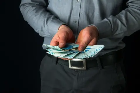 Mężczyzna w szarej koszuli trzyma w dłoniach polskie banknoty 50 i 100 zł.