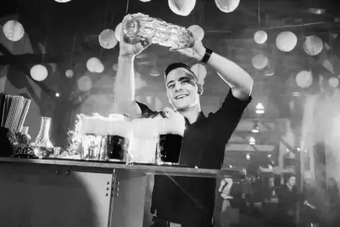 Uśmiechnięty barman z shakerem nad głową, przygotowuje drinki z dymem.