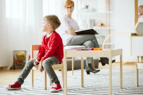 Pojke i röd tröja sitter på en stol under en autism utredning barn. En kvinna sitter bakom ett bord.