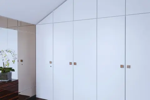 Armario empotrado con puertas blancas y tiradores cuadrados. Reflejos de orquídeas y un mueble de madera oscura en el espejo. Ideal para cambiar puertas armario empotrado.