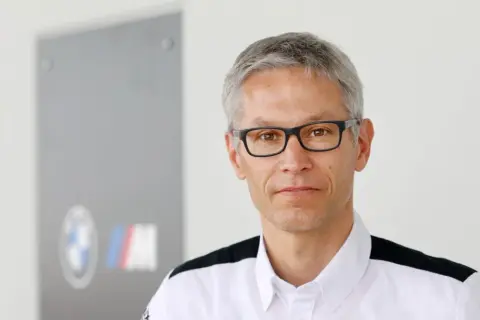 Mike Krack, con gafas y camisa blanca con detalles negros, posa frente a un logo de BMW M.