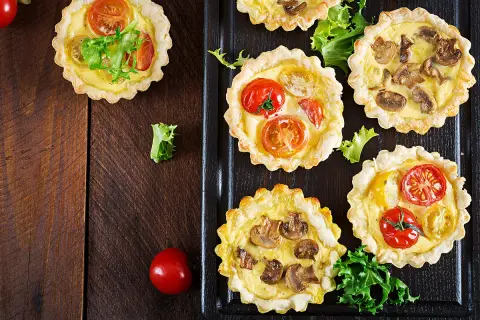 Mini quiches apéro aux tomates et champignons, garnies de feuilles de salade verte, sur fond de bois sombre.
