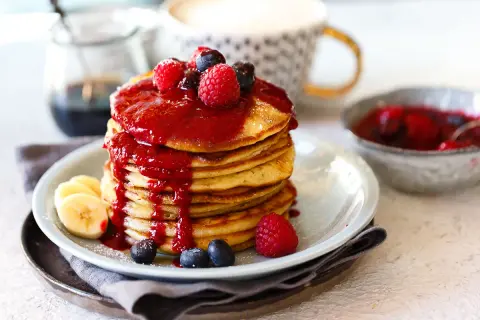 Ein Stapel goldbrauner Dinkel Pancakes, übergossen mit roter Beerensauce und garniert mit frischen Beeren und Bananenscheiben.