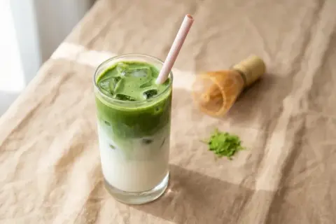 Un verre de pâte minute claire au matcha, glacé et rafraîchissant, avec une paille rose. Un fouet à matcha et une pincée de poudre verte à côté.