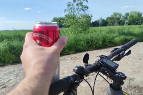 Rowerzysta trzyma puszkę napoju, planując jazdę rowerem po alkoholu. W tle zielone pola i błękitne niebo.