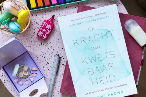 Boek 'De Kracht van Kwetsbaarheid' en oordopjes, ideaal voor wie last heeft van omgevingsgeluid. Creatieve spullen op een roze ondergrond.